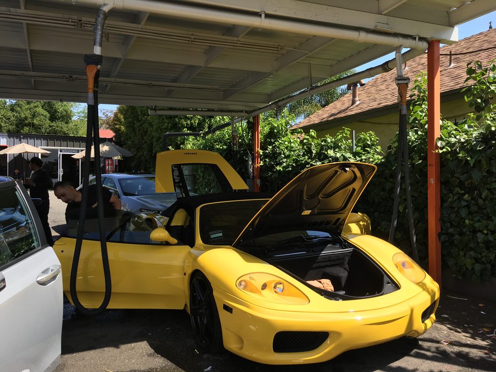A yellow sports car with its hood open under the orange canopy columns, a JN Car Wash crew member working on it.