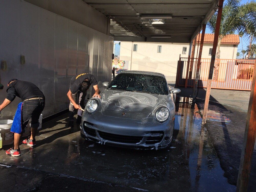 Two workers washing a silver Porsche covered in soap suds under the open canopy, water standing on the concrete.