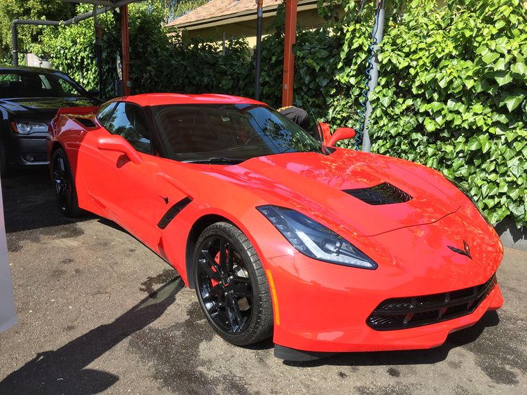 A red Corvette parked on the lot in front of a tall green hedge.